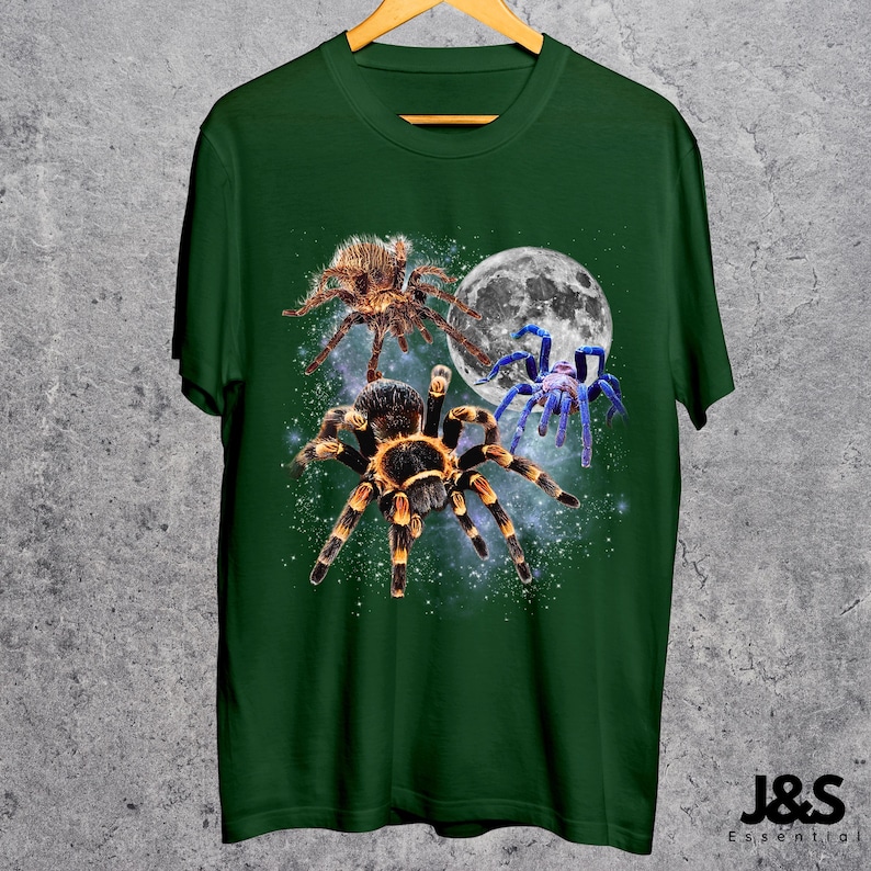 May include: Green t-shirt featuring a graphic of three spiders in front of a full moon and a starry background. The spiders are brown, black, and blue. The shirt has a crew neck and short sleeves. The brand name "J&S Essential" is at the bottom right.