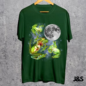 May include: Dark green t-shirt featuring a vibrant graphic of several frogs with a full moon in a starry night sky. The frogs are depicted in shades of green and yellow, with intricate details. The design is centered on the shirt.
