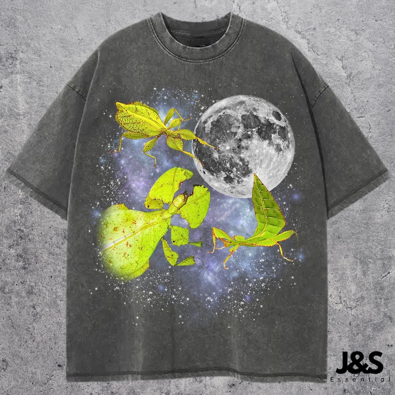 May include: Dark gray t-shirt featuring a graphic of a full moon, green leaf insects, and a starry galaxy background. The shirt has a crew neck and short sleeves. The text "J&S Essential" is at the bottom right.