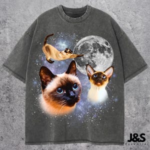 Puede incluir: Camiseta gris oscuro con un gráfico de tres gatos siameses con ojos azules sobre un fondo de luna llena y cielo estrellado. La camiseta tiene cuello redondo y mangas cortas. El texto "J&S Essential" está impreso en la parte inferior derecha.