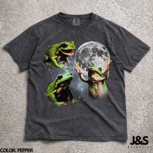 Può includere: T-shirt color pepe con un design stravagante di tre rane verdi e una luna piena sullo sfondo. Le rane sono in varie pose, con una che sembra allungare la mano verso la luna. La maglietta ha un aspetto vintage e lavato.