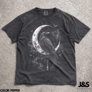 May include: A pepper-colored short-sleeve t-shirt featuring a white graphic print of a raven perched on a crescent moon. The design includes intricate spiderweb details, hanging crystal-like elements, small stars, and bats against a smoky background.