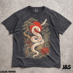 Könnte beinhalten: Ein pfefferfarbenes T-Shirt mit einem Grafikdruck einer weißen Schlange mit roten Mustern, die sich vor einer großen roten Sonne windet. Das Design zeigt einen fliegenden Kranich, einen Kiefernbaum, Bambus und rote sowie gelbe Blumen.