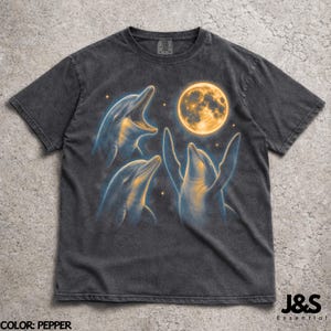 May include: Dark grey t-shirt with a graphic of three dolphins and a full moon. The dolphins are blue and yellow, with one leaping and the others looking up. The text "J&S Essential" is in the lower right corner. The color is "Pepper".