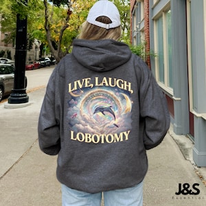 May include: Dark gray hoodie with the text "LIVE, LAUGH, LOBOTOMY" in a whimsical font. The back of the hoodie features a dolphin leaping through a rainbow-colored archway, surrounded by clouds and crescent moons. The hoodie has a drawstring hood.