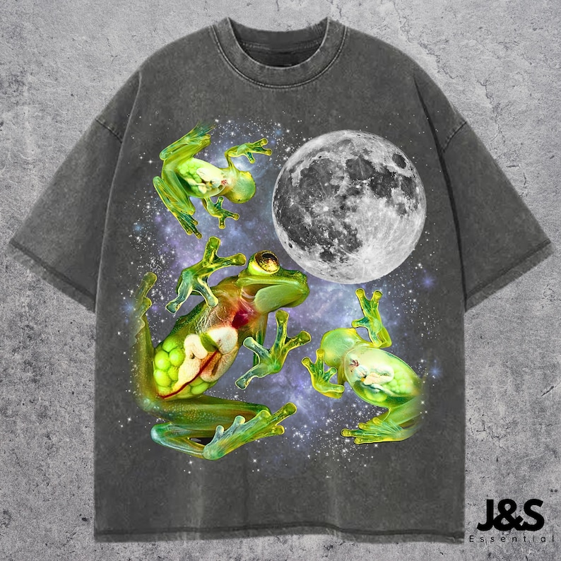 May include: Dark gray t-shirt featuring a vibrant graphic of three green frogs and a full moon against a starry background. The frogs have detailed, translucent bodies. The shirt has a relaxed fit and a crew neck.