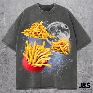 May include: Dark gray t-shirt featuring a graphic design of french fries and a full moon against a starry background. The fries are in a red container and scattered around the moon. The shirt has the text "J&S Essential" at the bottom.
