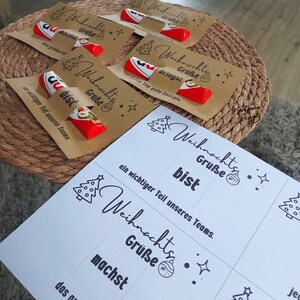 K&ouml;nnte beinhalten: Mehrere kleine Geschenke sind auf einer gewebten Oberfl&auml;che angeordnet. Jedes Geschenk enth&auml;lt einen rot-wei&szlig;en Schokoriegel, der an einer braunen Karte mit den Worten "Weihnachts Gr&uuml;&szlig;e" und einer Weihnachtsbaum-Illustration befestigt ist. Ein wei&szlig;es Blatt mit demselben Text ist ebenfalls vorhanden.