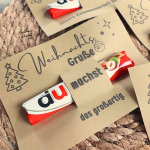 K&ouml;nnte beinhalten: Kraftpapierkarten mit Weihnachtsgr&uuml;&szlig;en und einem Kinder Bueno Schokoriegel. Die Karten haben ein Weihnachtsbaumdesign und die Worte "Weihnachts Gr&uuml;&szlig;e" und "machst das gro&szlig;artig" in Schwarz gedruckt.