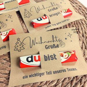 K&ouml;nnte beinhalten: Karten aus Kraftpapier mit Weihnachtsgr&uuml;&szlig;en und Schokoriegeln. Die Karten zeigen eine Weihnachtsbaum-Illustration und die Worte "Weihnachts Gr&uuml;&szlig;e" und "du bist". Die Schokoriegel sind rot, wei&szlig; und schwarz.