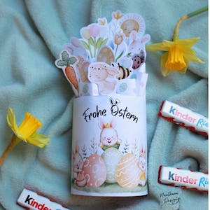 May include: A white cylindrical Easter-themed paper craft with the words "Frohe Ostern" and illustrations of bunnies, eggs, and flowers. The top of the craft features additional bunny and floral cutouts. Two yellow daffodils and two chocolate bars are also in the image.
