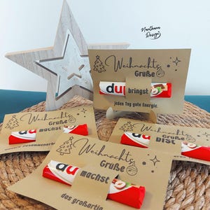 K&ouml;nnte beinhalten: Weihnachtsgru&szlig;karten aus Kraftpapier mit einem Kinder Bueno Schokoriegel. Die Karten haben ein Weihnachtsbaum- und Sterndesign mit deutschem Text. Im Hintergrund befindet sich ein Holzstern.