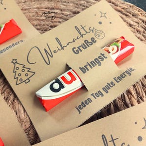 K&ouml;nnte beinhalten: Eine Karte aus Kraftpapier mit Weihnachtsgr&uuml;&szlig;en und einer Weihnachtsbaum-Illustration. Ein rot-wei&szlig;es Schokoriegel steckt teilweise in einem Schlitz der Karte. Der Text "bringst jeden Tag gute Energie" ist ebenfalls sichtbar.