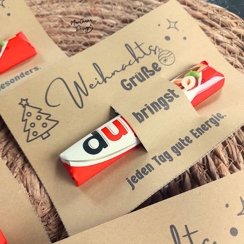 K&ouml;nnte beinhalten: Eine Karte aus Kraftpapier mit Weihnachtsgr&uuml;&szlig;en und einer Weihnachtsbaum-Illustration. Ein rot-wei&szlig;es Schokoriegel steckt teilweise in einem Schlitz der Karte. Der Text "bringst jeden Tag gute Energie" ist ebenfalls sichtbar.