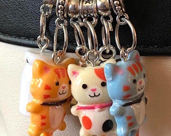 May include: A silver charm bracelet with three cat charms. The charms are orange, white, and blue, each with a cute cat design. The orange cat has orange stripes, the white cat has red spots, and the blue cat has blue stripes.