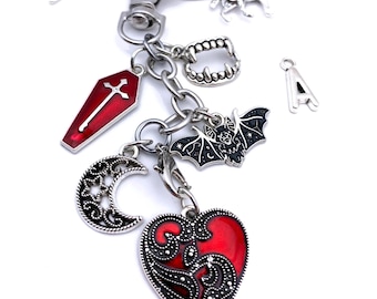 May include: Silver-toned keychain with various charms, including a red coffin, a crescent moon, a heart, a bat, and a set of fangs. The heart charm has a red center with a decorative silver border. Additional charms include a letter "A" and two mythical creatures.