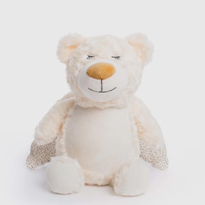 May include: A cream-coloured plush teddy bear with a light brown nose and closed eyes. The bear has soft, textured fur and small black polka dots on the inner ears and paws. The bear is sitting upright.