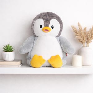 May include: A soft penguin toy with a grey head and wings, a white belly, and yellow feet. The penguin is on a white shelf with a small plant, books, a candle, and a vase with decorative grass.