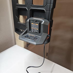 Könnte beinhalten: Ein schwarzes und graues RIDGID-Batterieladegerät mit orangefarbenem Akzent, montiert an einem schwarzen Kunststoffhalter. Das Ladegerät hat ein Warnschild und ein schwarzes Netzkabel. Das Ladegerät befindet sich auf einer weißen Oberfläche.