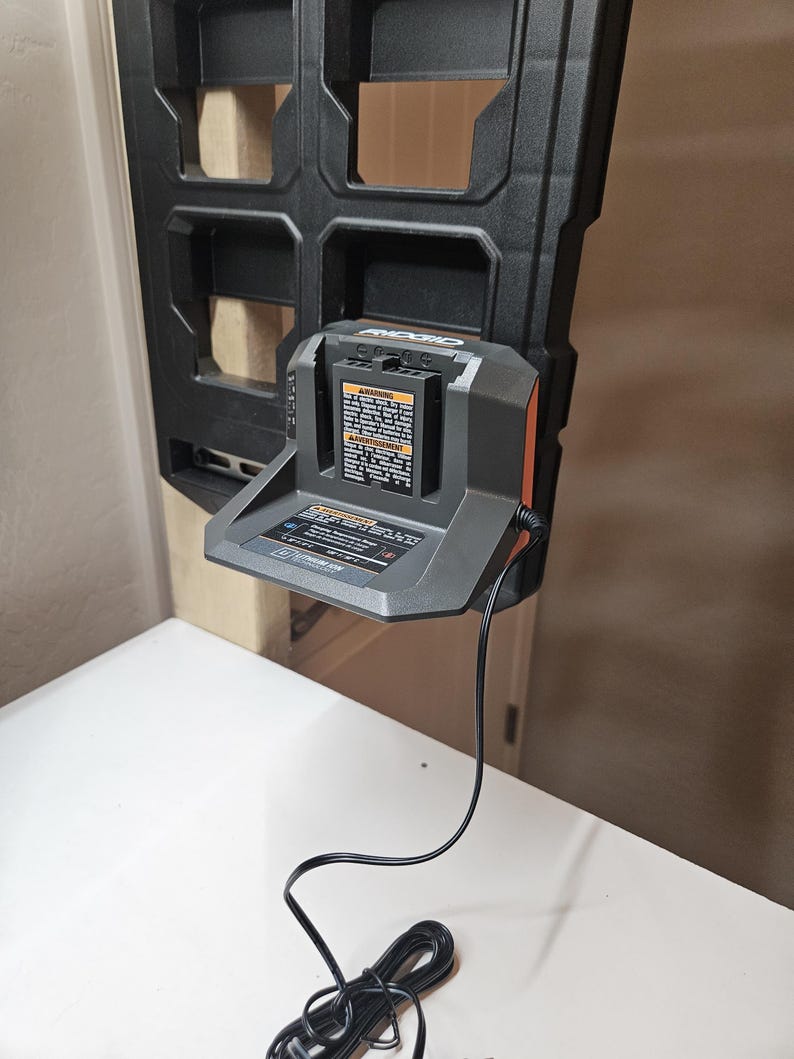 May include: A black and gray RIDGID battery charger with an orange accent, mounted on a black plastic holder. The charger has a warning label and a black power cord. The charger is on a white surface.
