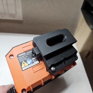 May include: An orange and black power tool battery with a black 3D-printed accessory attached. The battery has a label with text and a safety symbol. The accessory appears to be a holder or mount.