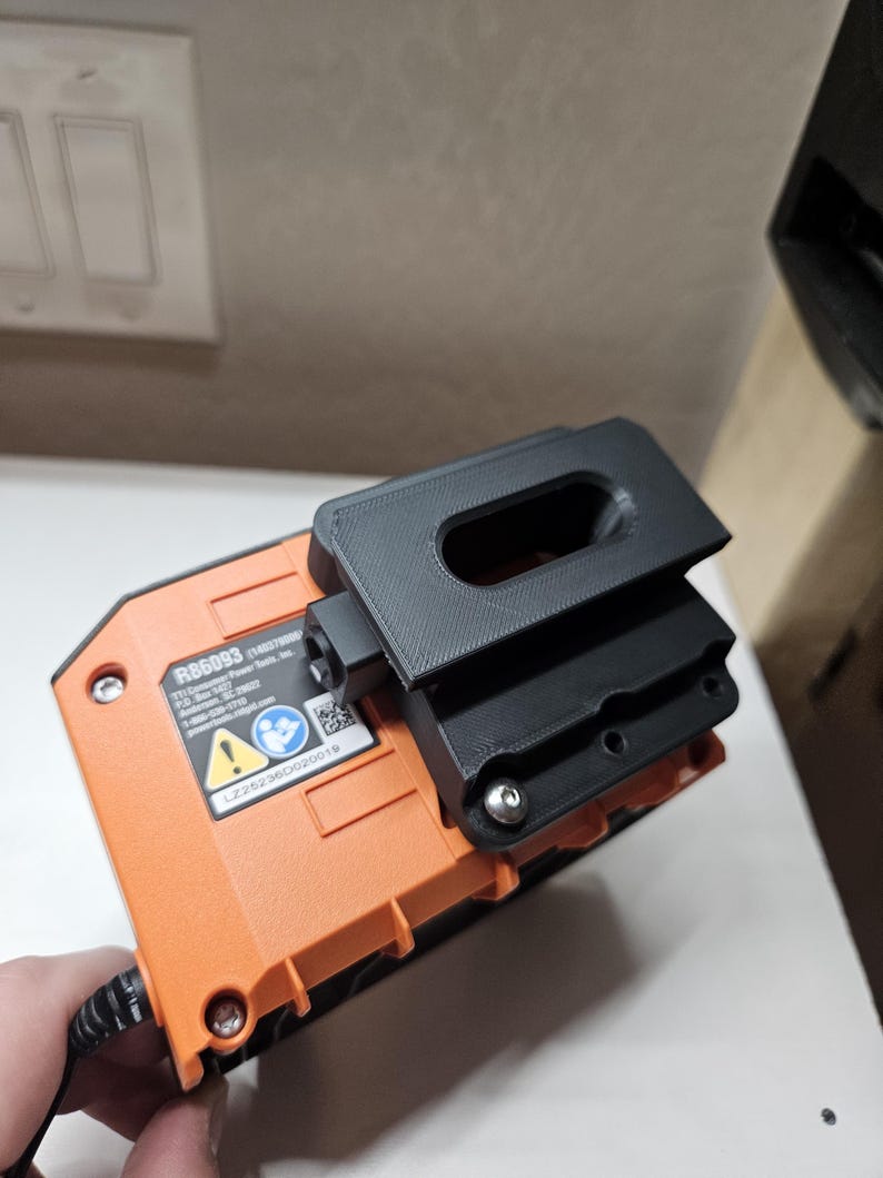May include: An orange and black power tool battery with a black 3D-printed accessory attached. The battery has a label with text and a safety symbol. The accessory appears to be a holder or mount.