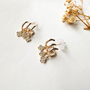 May include: Gold hoop earrings with three dangling charms, each charm is a small flower with clear rhinestones.
