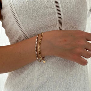 Op de afbeelding: Twee gouden armbanden om een pols. De ene armband is een delicate ketting met kleine, heldere stenen. De andere armband is een dikkere ketting met een kleine ronde hanger.