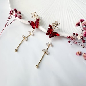Puede incluir: Pendientes dorados con diseño de mariposa roja y detalles de cristal transparente. Los pendientes tienen un elemento floral en la parte superior y un tallo colgante con detalles de cristal. Sobre un fondo blanco.