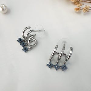 May include: Silver hoop earrings with three hoops and a dangling charm of three blue crystal flower designs.