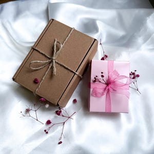 K&ouml;nnte beinhalten: Zwei Geschenkboxen auf einer wei&szlig;en Satin-Oberfl&auml;che. Eine ist braun, mit Bindfaden gebunden. Die andere ist rosa, mit einem rosa Band umwickelt und mit kleinen Blumen verziert. Die Boxen sind von Zweigen mit winzigen roten Bl&uuml;ten umgeben.