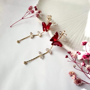 May include: Gold-coloured butterfly earrings with red jewel wings and clear crystal details. The earrings have a floral stud and a delicate stem design with crystal leaves. The earrings are displayed on a white surface with pink flowers.