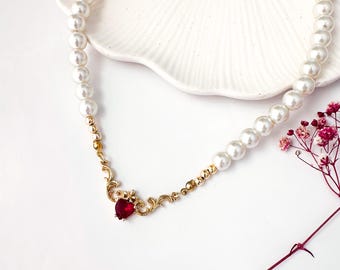 Puede incluir: Un delicado collar de oro con un colgante de corazón rojo y cuentas de perlas blancas.