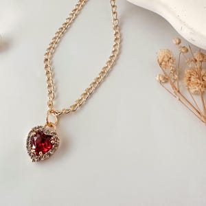 Peut inclure: Un collier en chaîne dorée avec un pendentif en forme de cœur rouge. Le cœur est entouré de strass transparents.