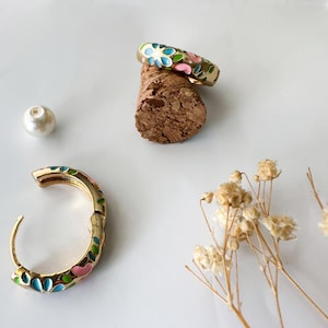 K&ouml;nnte beinhalten: Goldfarbenes Schmuckset mit einem Ring und einem Creolen-Ohrring, beide mit einem Blumenmuster aus blauem, rosa und gr&uuml;nem Emaille verziert. Der Ring liegt auf einem Korken, w&auml;hrend der Ohrring offen ist und das Design zeigt. Eine Perle und Trockenblumen vervollst&auml;ndigen die Komposition.