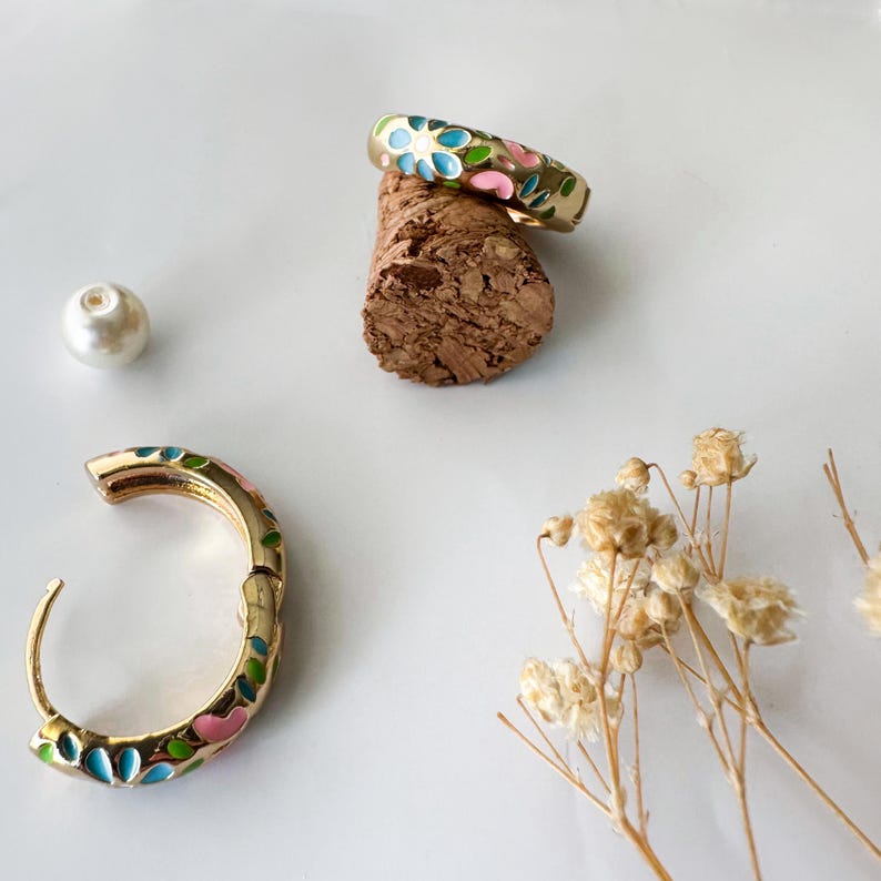 K&ouml;nnte beinhalten: Goldfarbenes Schmuckset mit einem Ring und einem Creolen-Ohrring, beide mit einem Blumenmuster aus blauem, rosa und gr&uuml;nem Emaille verziert. Der Ring liegt auf einem Korken, w&auml;hrend der Ohrring offen ist und das Design zeigt. Eine Perle und Trockenblumen vervollst&auml;ndigen die Komposition.