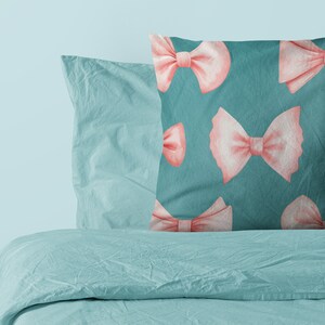 K&ouml;nnte beinhalten: Ein blaues Kissen mit einem Muster aus rosa Schleifen. Das Kissen liegt auf einem Bett mit einem blauen Bettbezug.