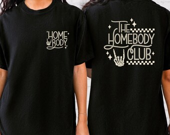 May include: Two black t-shirts with white text. One shirt says "Homebody" with a rock and roll hand symbol. The other shirt says "The Homebody Club" with a rock and roll hand symbol and a checkered pattern.