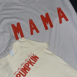 Pode incluir: Duas camisetas, uma cinza claro com a palavra "MAMA" em vermelho, e uma creme com o texto "mama's pumpkin" em vermelho.