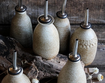 May include: A collection of six ceramic oil bottles with stainless steel spouts. The bottles are various shades of beige and brown with speckled textures. Some bottles have a smooth surface, while others have a textured, carved design. The bottles are arranged on a wooden surface.