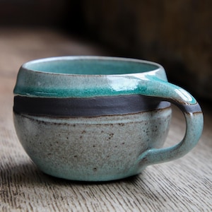 Może przedstawiać: Ręcznie robiony kubek ceramiczny z turkusową glazurą i ciemnoszarym paskiem wokół krawędzi. Kubek ma wygląd rustykalny i nakrapiany.
