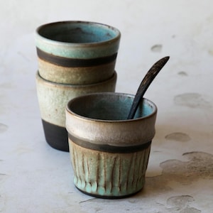 Może przedstawiać: Cztery ręcznie robione ceramiczne kubki. Trzy są ułożone w stos, a jeden z przodu. Kubki mają turkusowe wnętrze, ciemnobrązowy pasek i teksturowany, jasnozielony wygląd zewnętrzny. Ciemna drewniana łyżka spoczywa w przednim kubku.