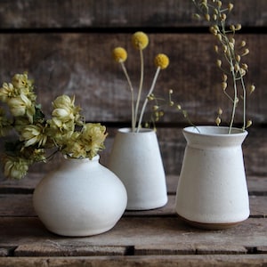 May include: Three white ceramic vases of different shapes and sizes. The vases are sitting on a wooden surface. The vase on the left is round and has a wide opening. The vase in the middle is tall and narrow. The vase on the right is short and wide.