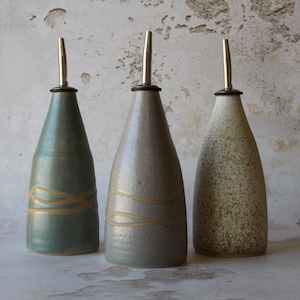 May include: Three ceramic oil bottles with stainless steel spouts. The bottles are in shades of green, grey, and beige. Each bottle has a unique texture and a decorative, light-coloured swirl design. The spouts are long and slender.