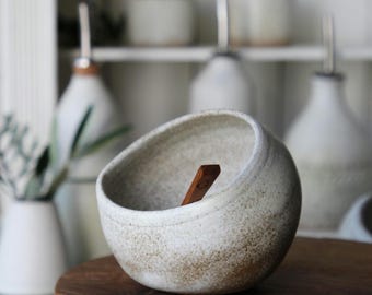 Peut inclure: Un bol en céramique blanc cassé de forme ronde avec une finition texturée et mouchetée et un bord incliné. Une petite cuillère en bois brun foncé repose à l'intérieur du bol, appuyée contre la paroi. Le bol présente un aspect rustique.