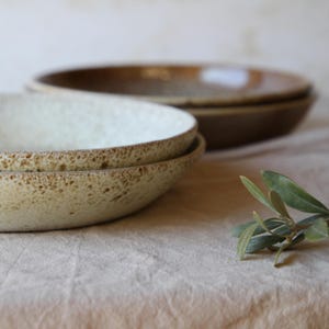 May include: Two ceramic bowls with a speckled, off-white interior and a brown rim are stacked in the foreground. Two more bowls are out of focus in the background. A sprig of green leaves is in the foreground. The bowls have a rustic, handmade appearance.