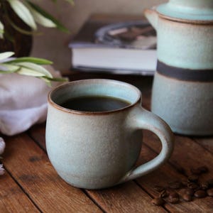 May include: A rounded ceramic mug with a matte, speckled light blue and beige glaze, featuring a dark brown rim and a curved handle. The mug is filled with dark coffee, showcasing a textured surface and a sturdy, thick-walled construction.