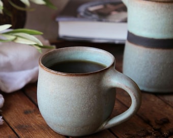 May include: A rounded ceramic mug with a matte, speckled light blue and beige glaze, featuring a dark brown rim and a curved handle. The mug is filled with dark coffee, showcasing a textured surface and a sturdy, thick-walled construction.