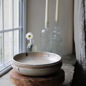 Puede incluir: Dos cuencos de cerámica apilados, con un esmalte moteado, colocados sobre un pequeño soporte de madera. Dos velas largas están en botellas de vidrio en el fondo, cerca de una ventana.