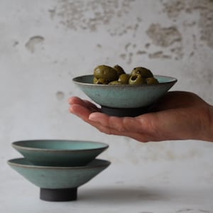 May include: A set of three teal ceramic bowls with a dark grey base. The bowls are stacked on top of each other. The top bowl is filled with green olives.